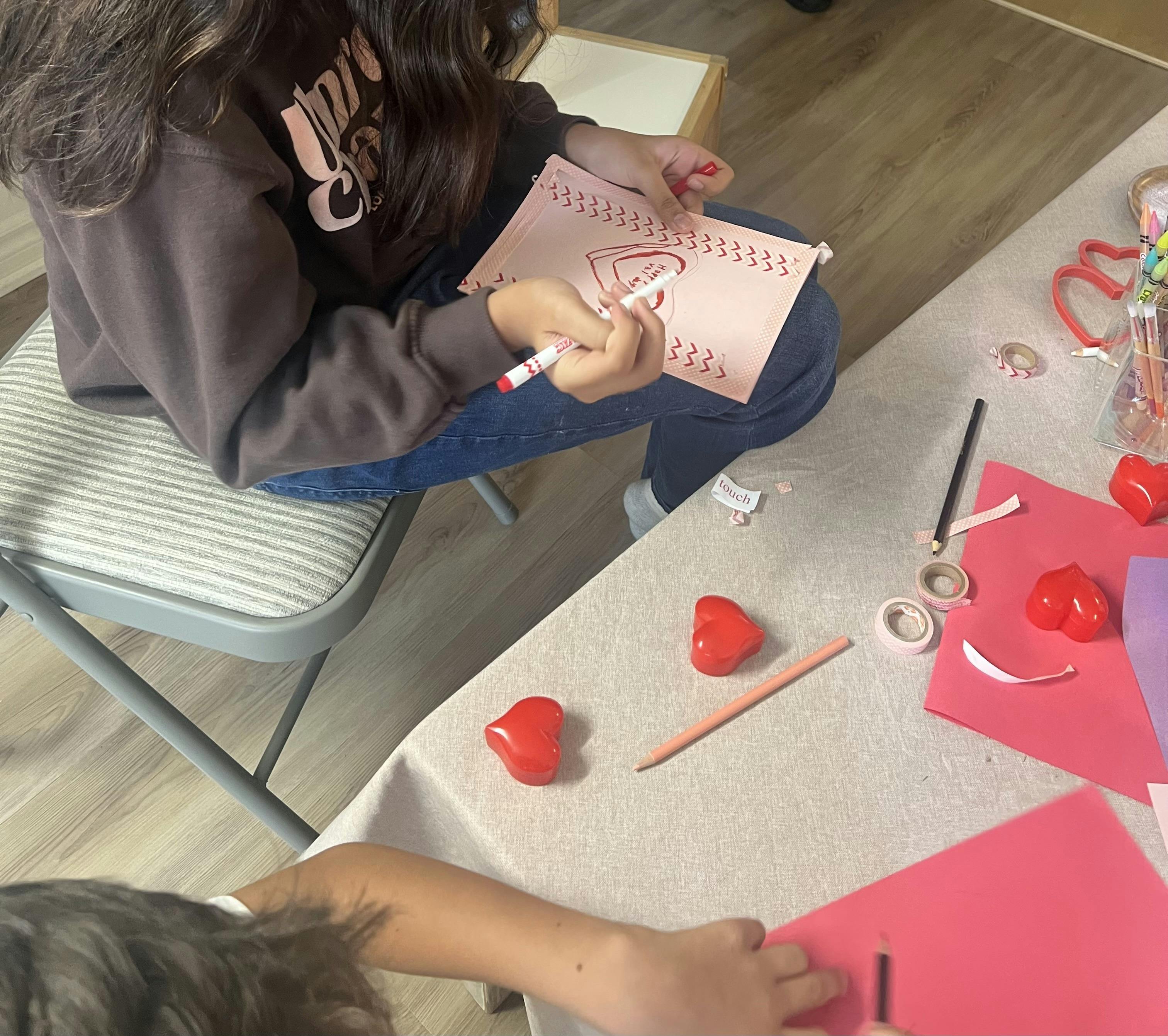 Person's hands holding a pink card with red marker art, making a heart drawing