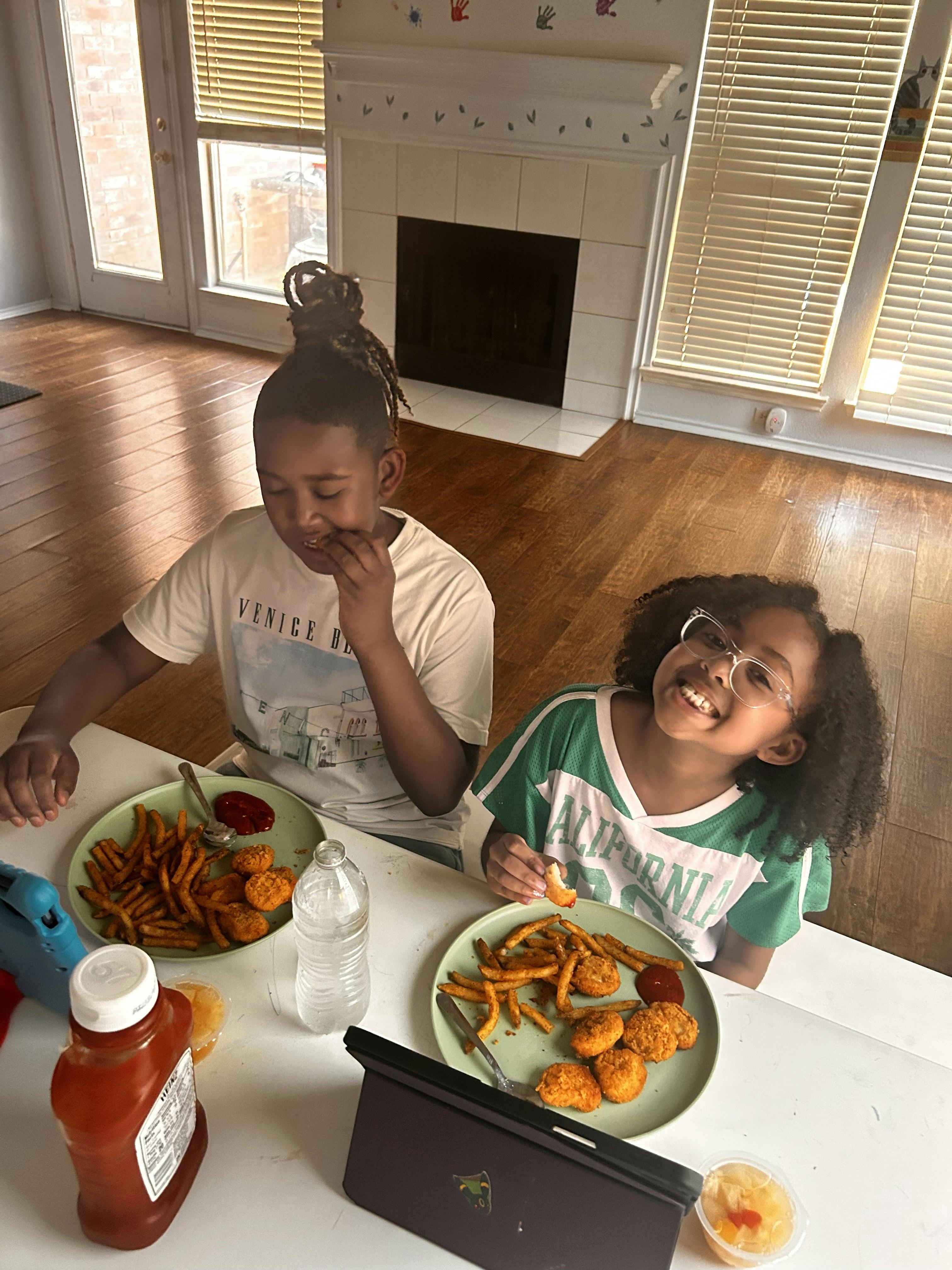 Two children sitting at a table eating chicken nuggets and fries