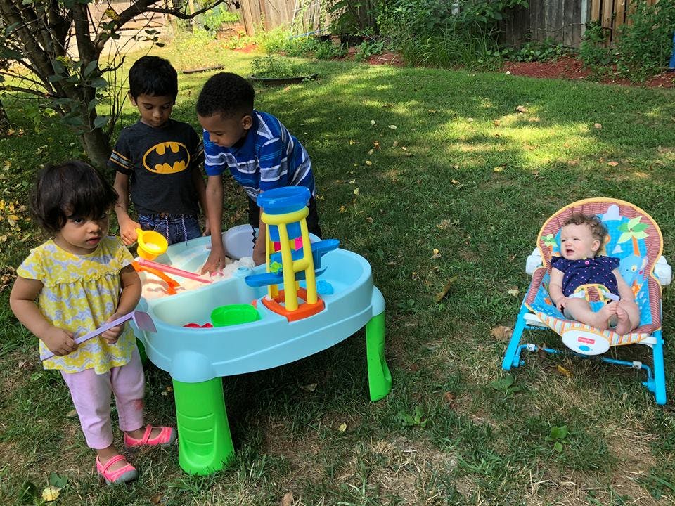 Three children playing with a water table outdoors, one baby in a bouncer