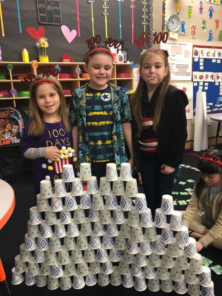 Three children standing behind a large pyramid of paper cups in a classroom