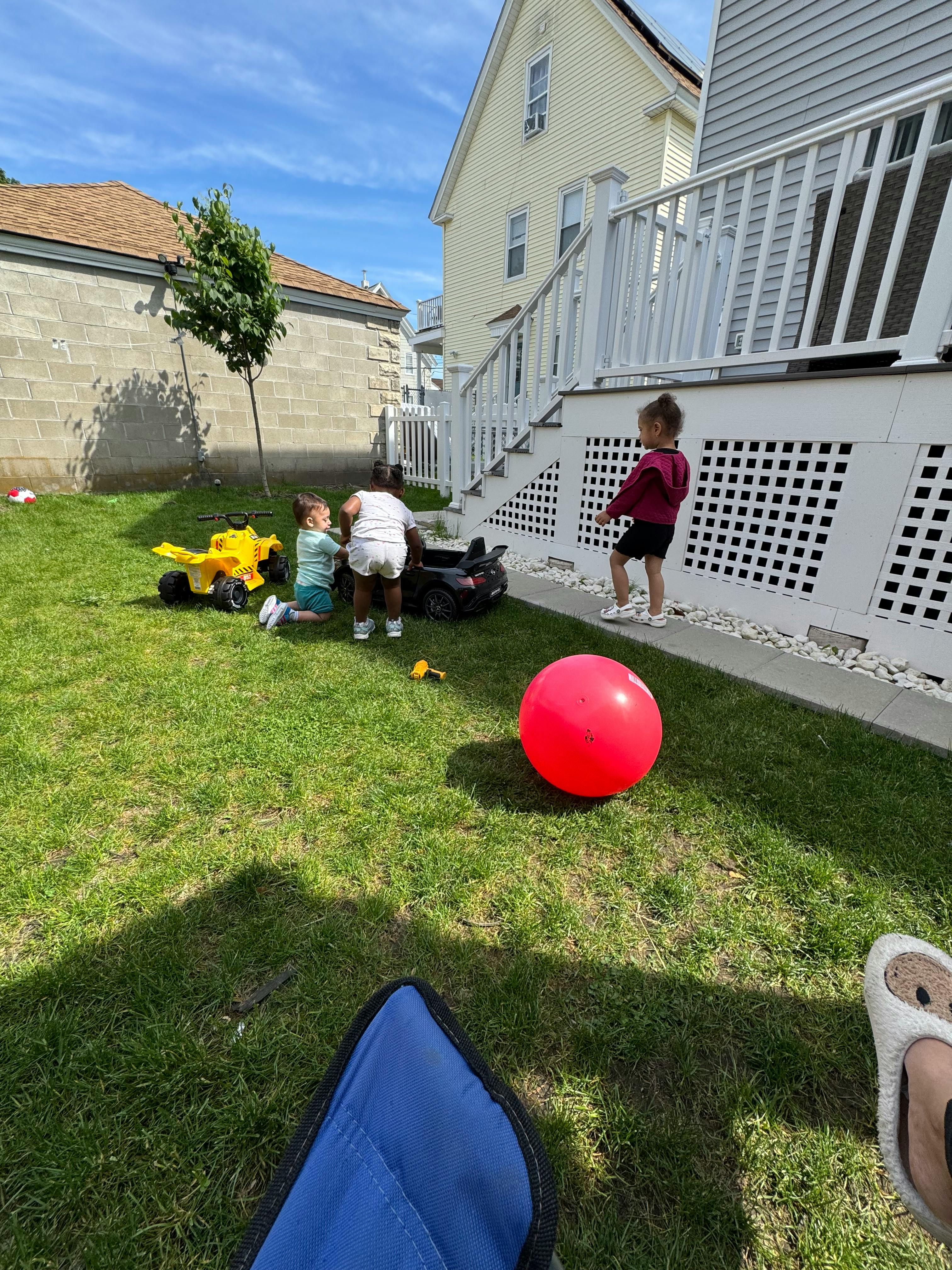 Three children playing in a grassy backyard with toys and near toys