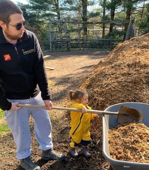 Person and child holding a shovel over a wheelbarrow full of wood chips