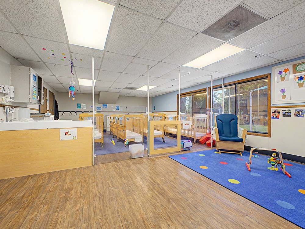 cribs in a daycare room with a rocking chair and play mat