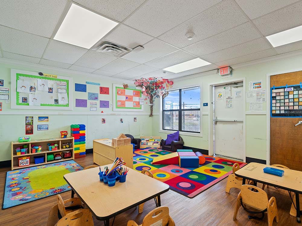 Colorful classroom with small tables and chairs, shelves with toys, and activity rugs