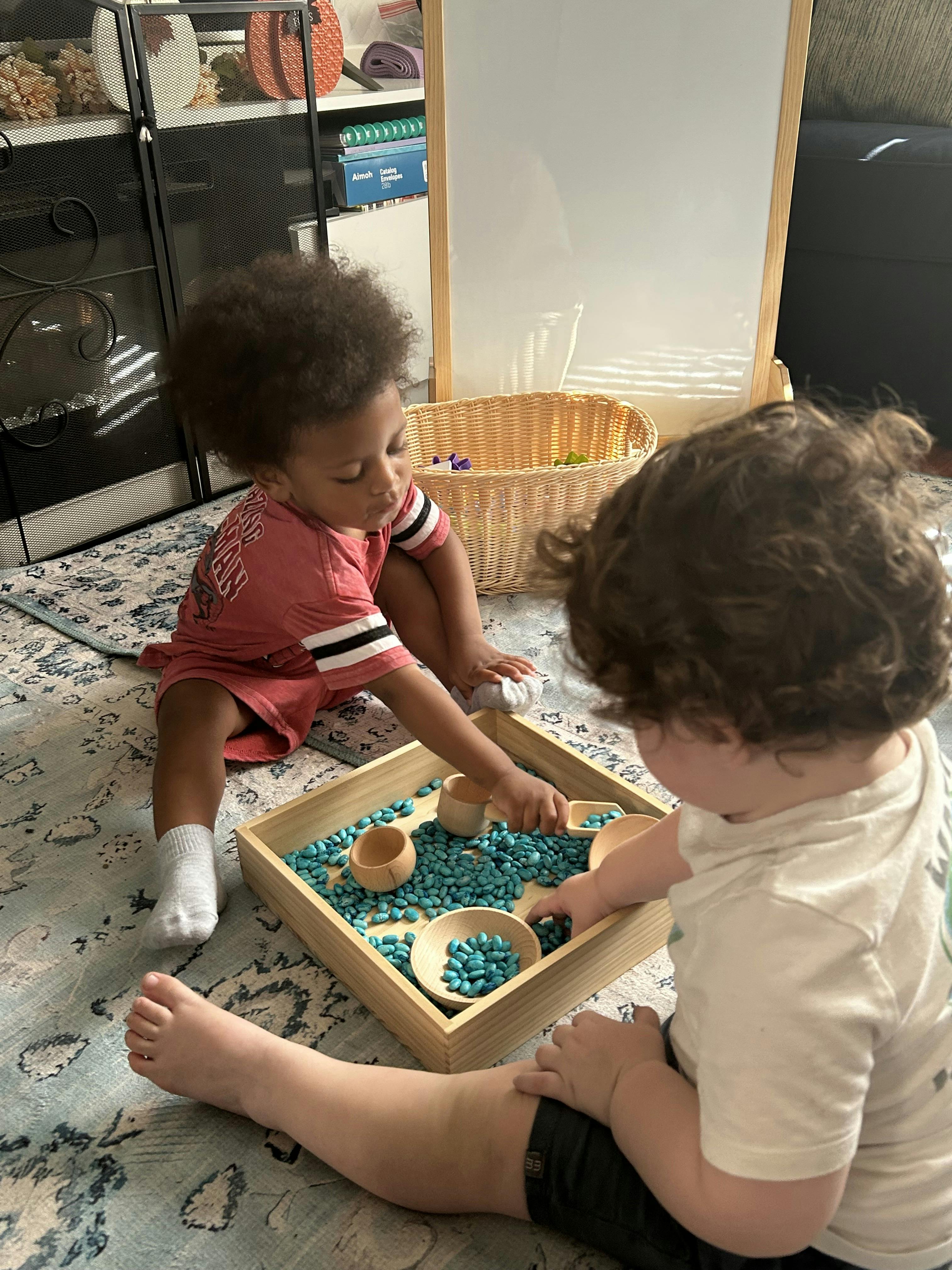 Two children playing with blue sensory beans in a wooden tray on a rug