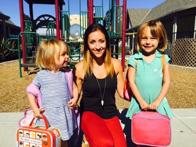 Person with two children holding lunchboxes in front of a playground