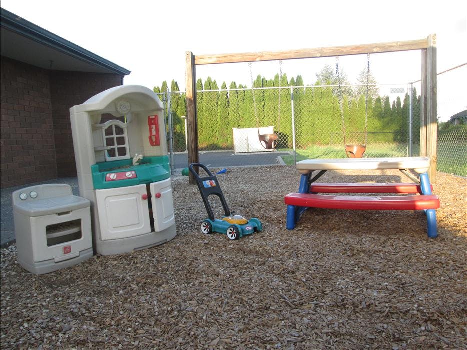 A play kitchen, a toy lawnmower, a picnic table, and swings on a woodchip ground
