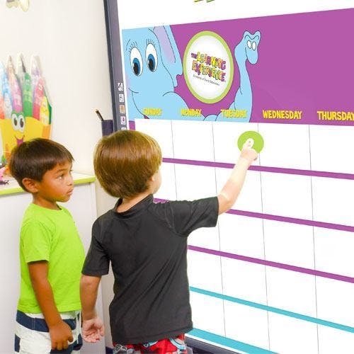 Child placing a green circle on a large calendar while another child watches