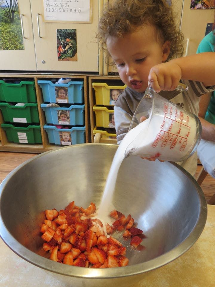Child pouring sugar from a measuring cup into a bowl of chopped strawberries