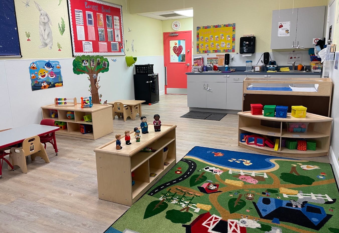 Classroom with small tables, shelves of toys, a play rug, and a changing station