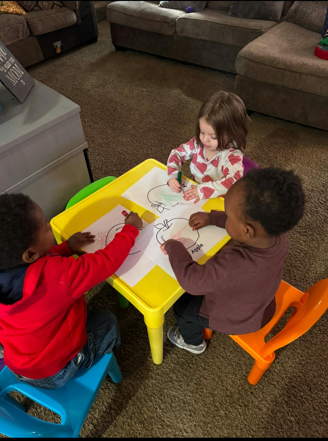 Three children sitting at a yellow table coloring drawings of apples