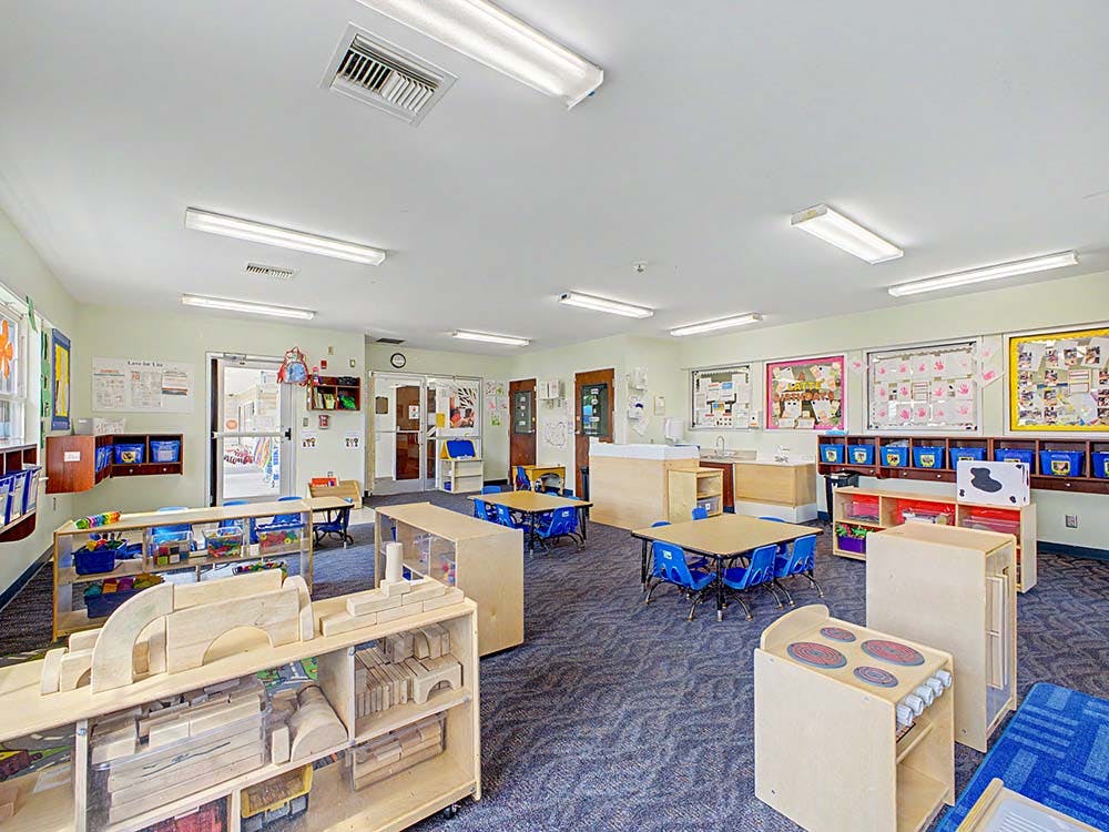 A brightly lit classroom with small tables and chairs, and wooden toy shelves
