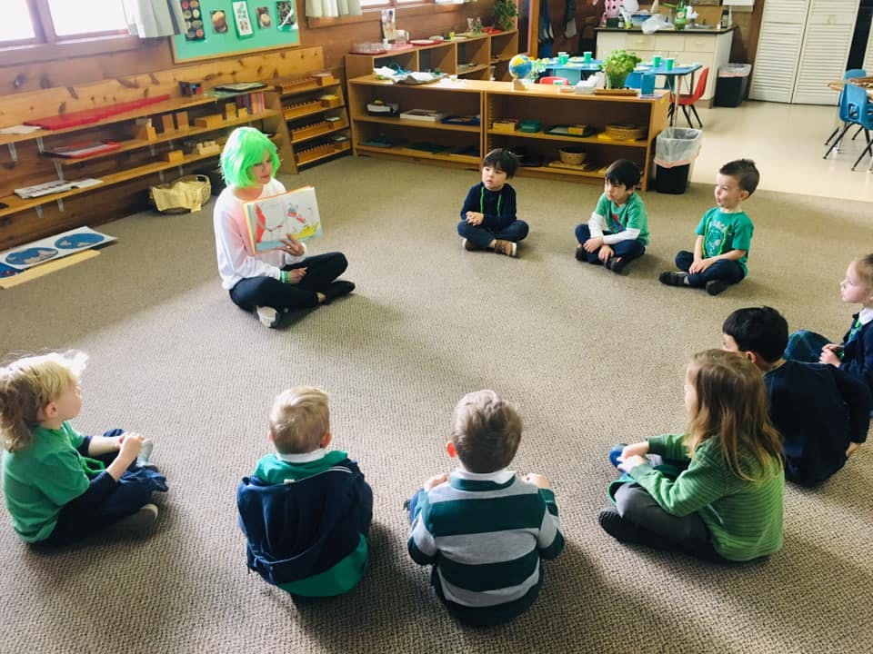 Person with green hair reading a book to children in a classroom