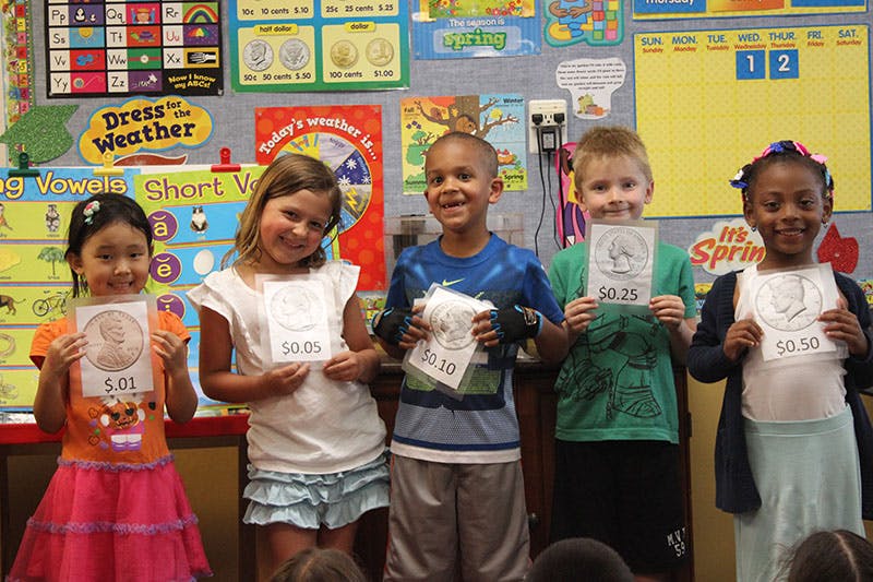 Five children holding up papers with drawings and numbers in a classroom