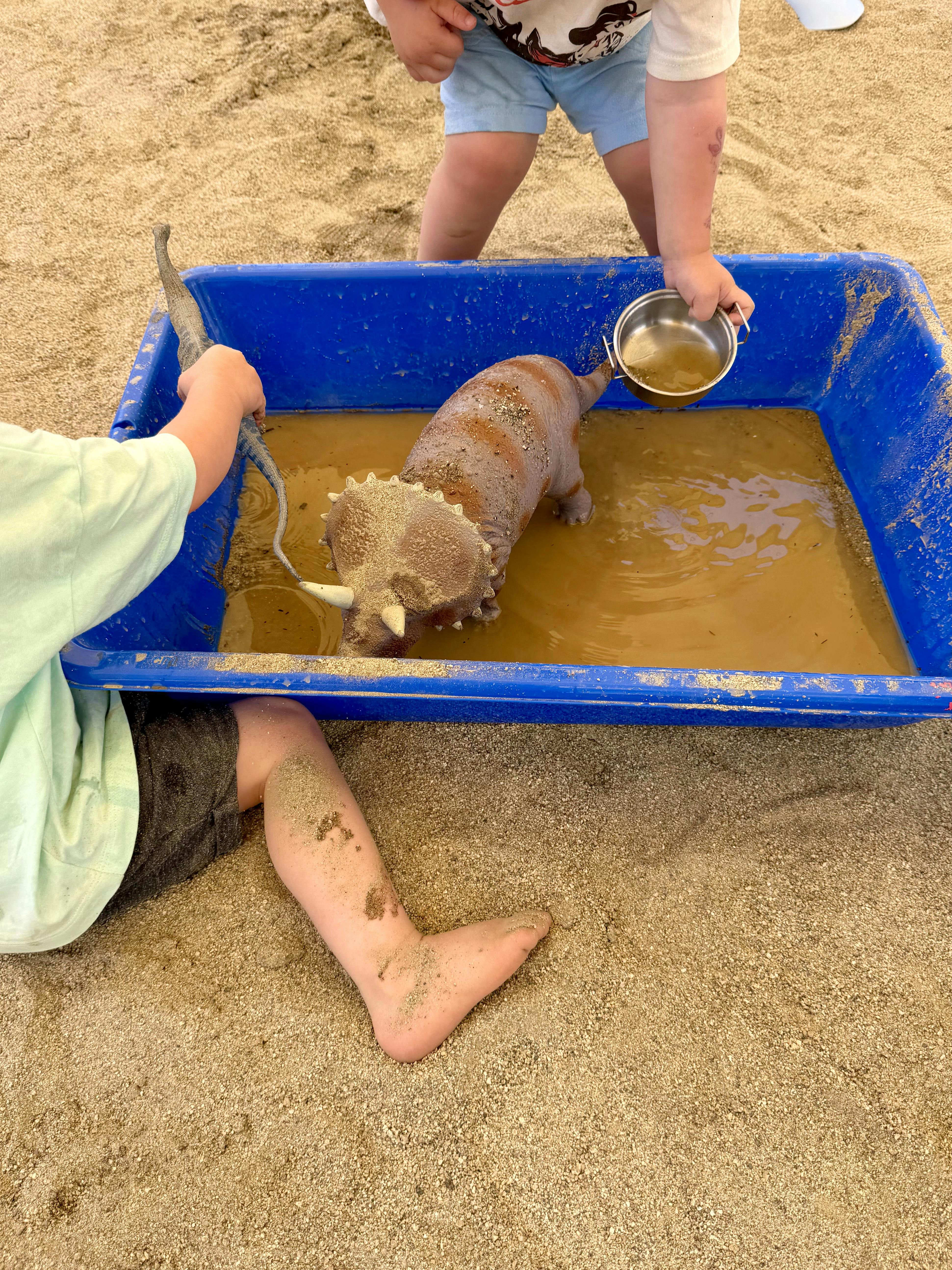 Children playing with a toy dinosaur in a blue bin of muddy water