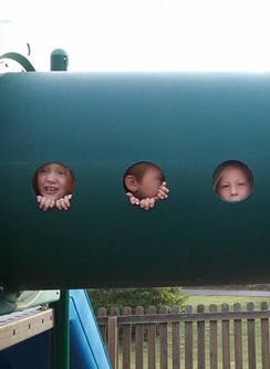 Three children looking out of holes in a green tunnel on a playground