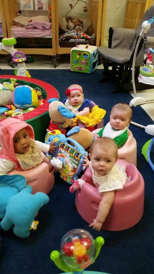 Four babies sitting in infant seats on a blue carpet with toys