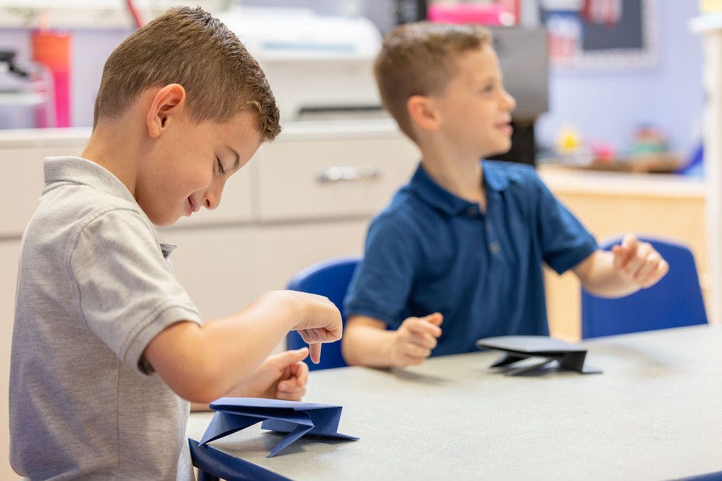 Child at a table looking down at an object, another child in background