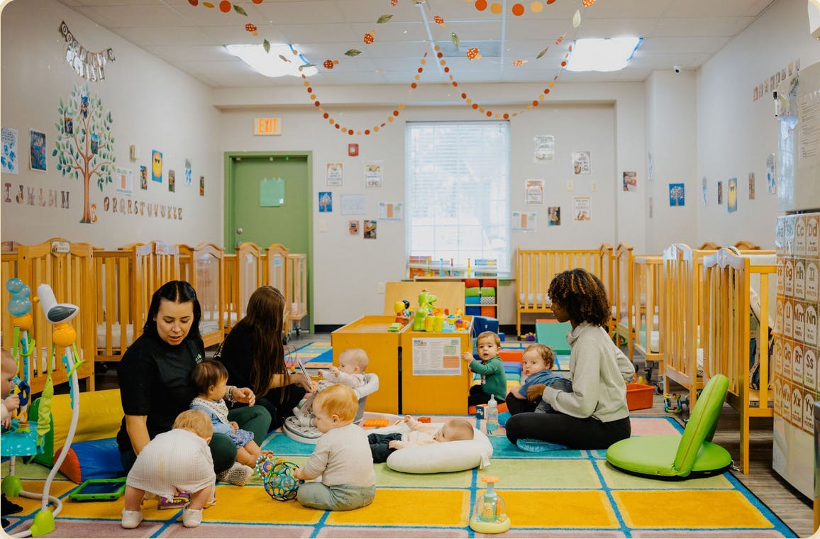 People with babies and infants on a colorful carpeted floor in a room with cribs