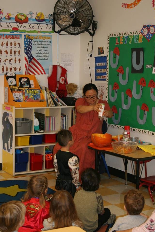 Person pouring liquid into a pumpkin while children watch in a classroom