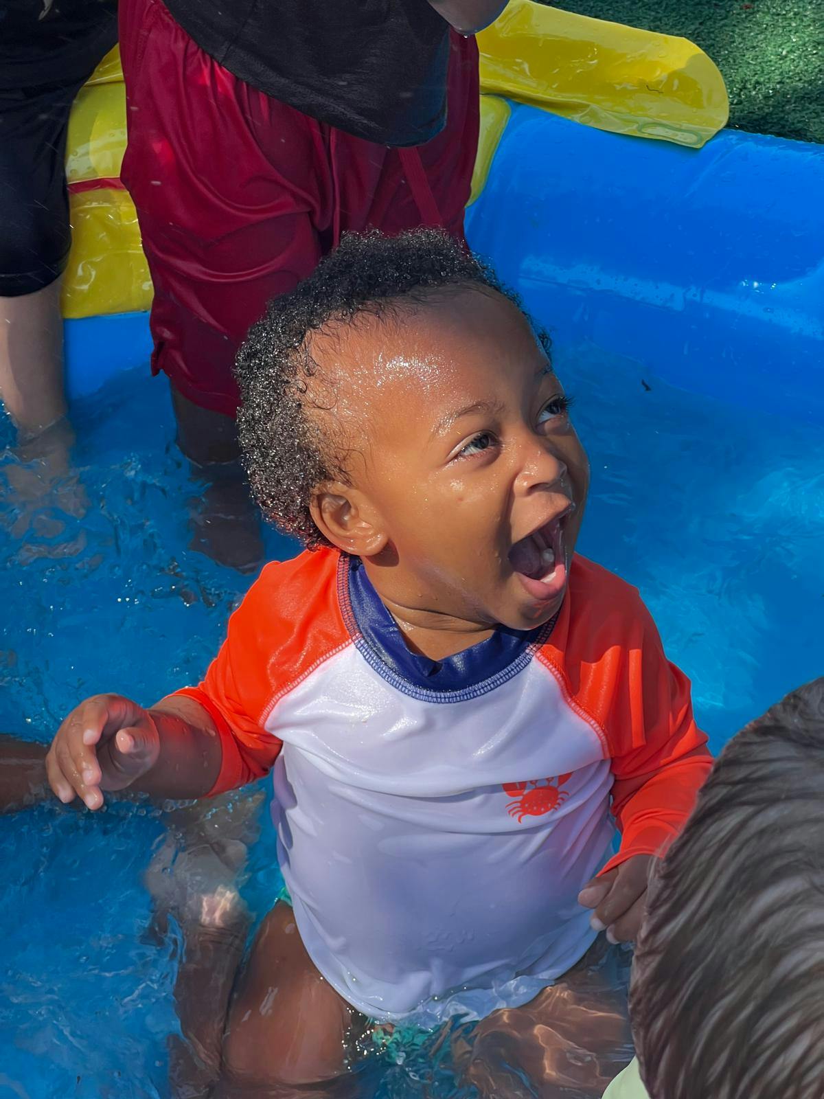Child in a blue pool wearing a white and orange swim shirt