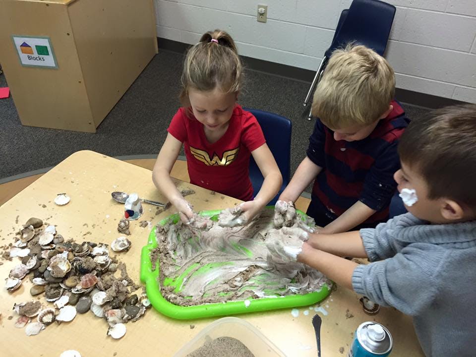 Three children playing with mud and shells at a table