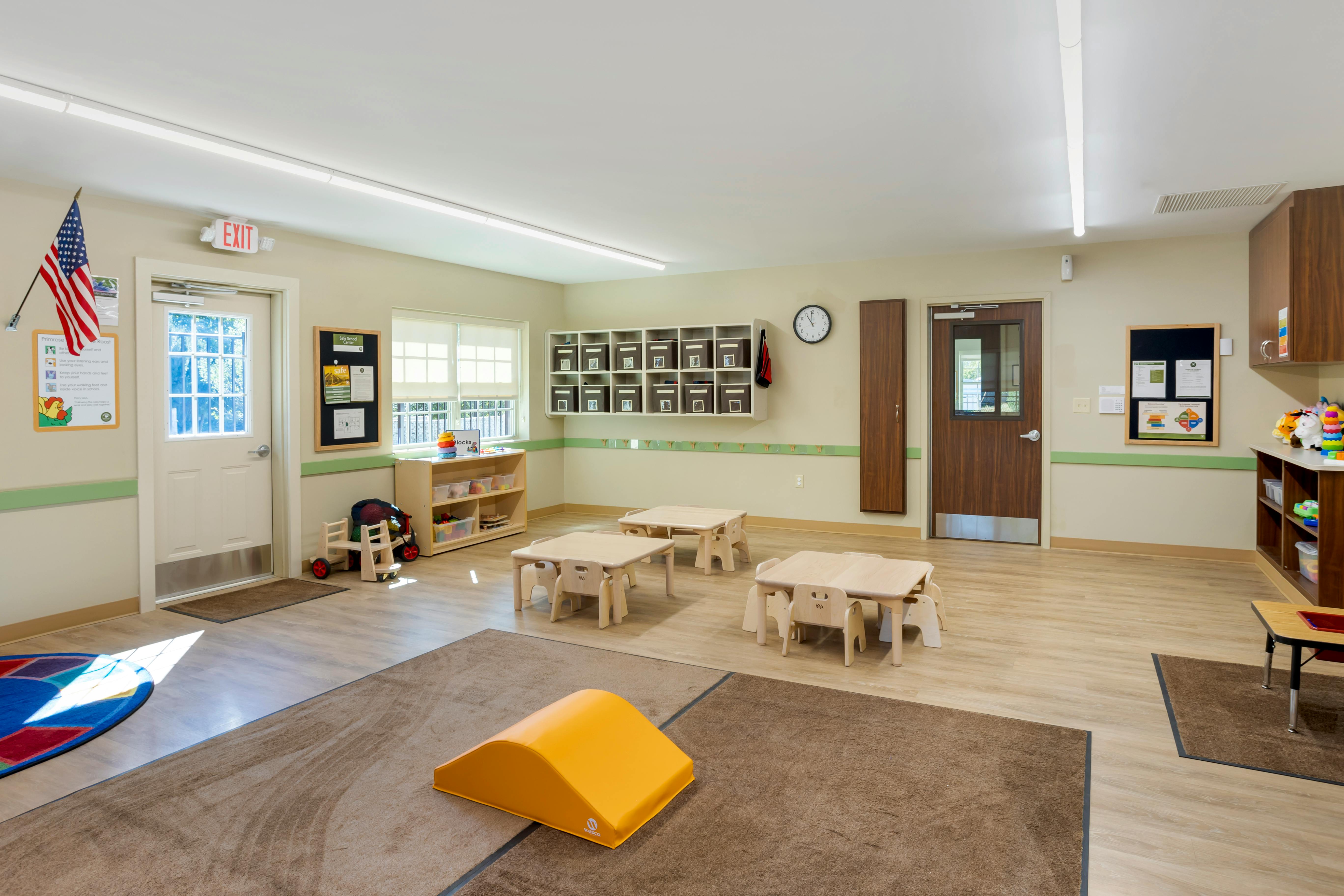 Classroom with small tables and chairs, shelves with bins, and an exit sign