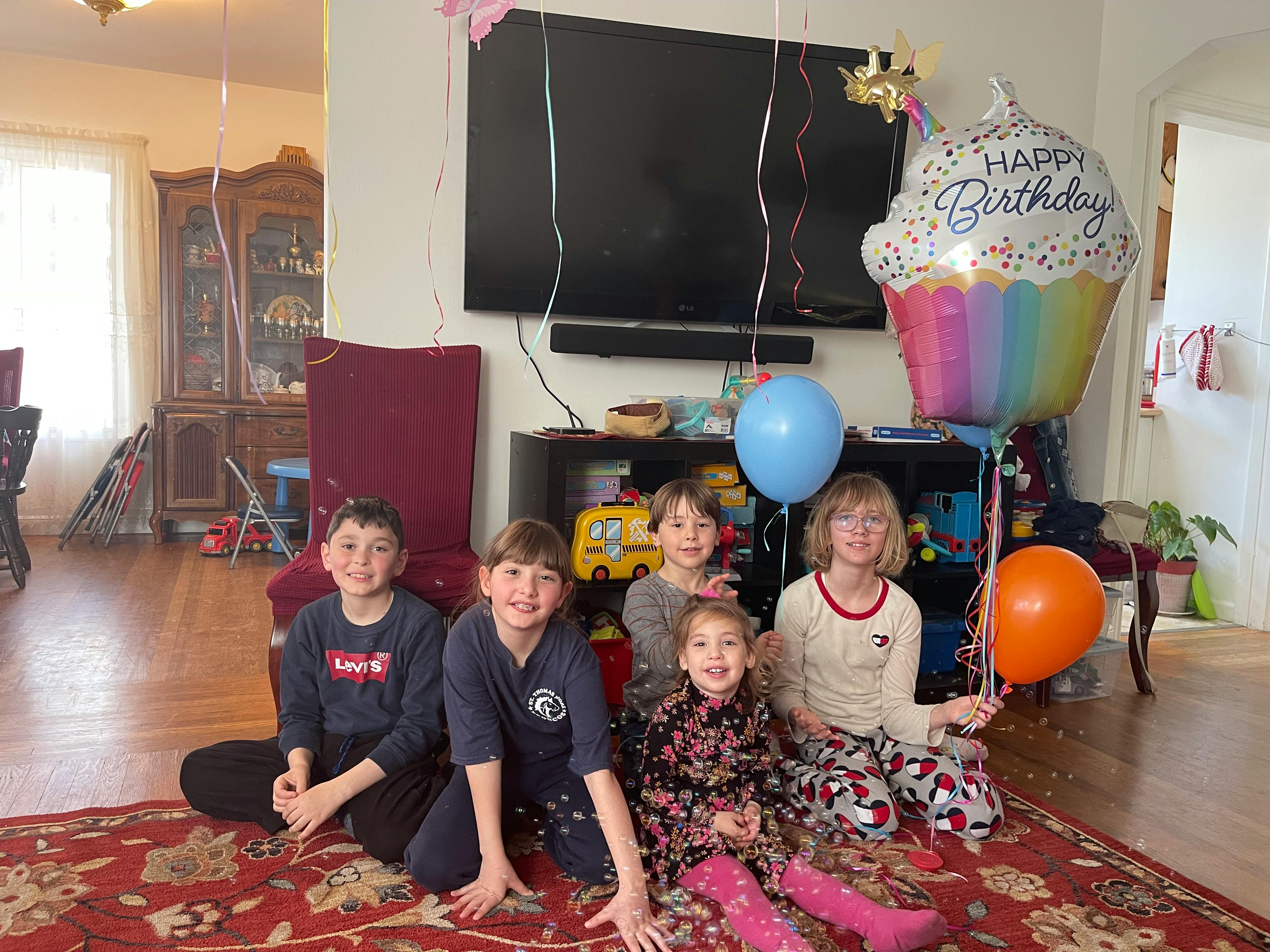 Five children sitting on a rug with balloons and a Happy Birthday cupcake balloon