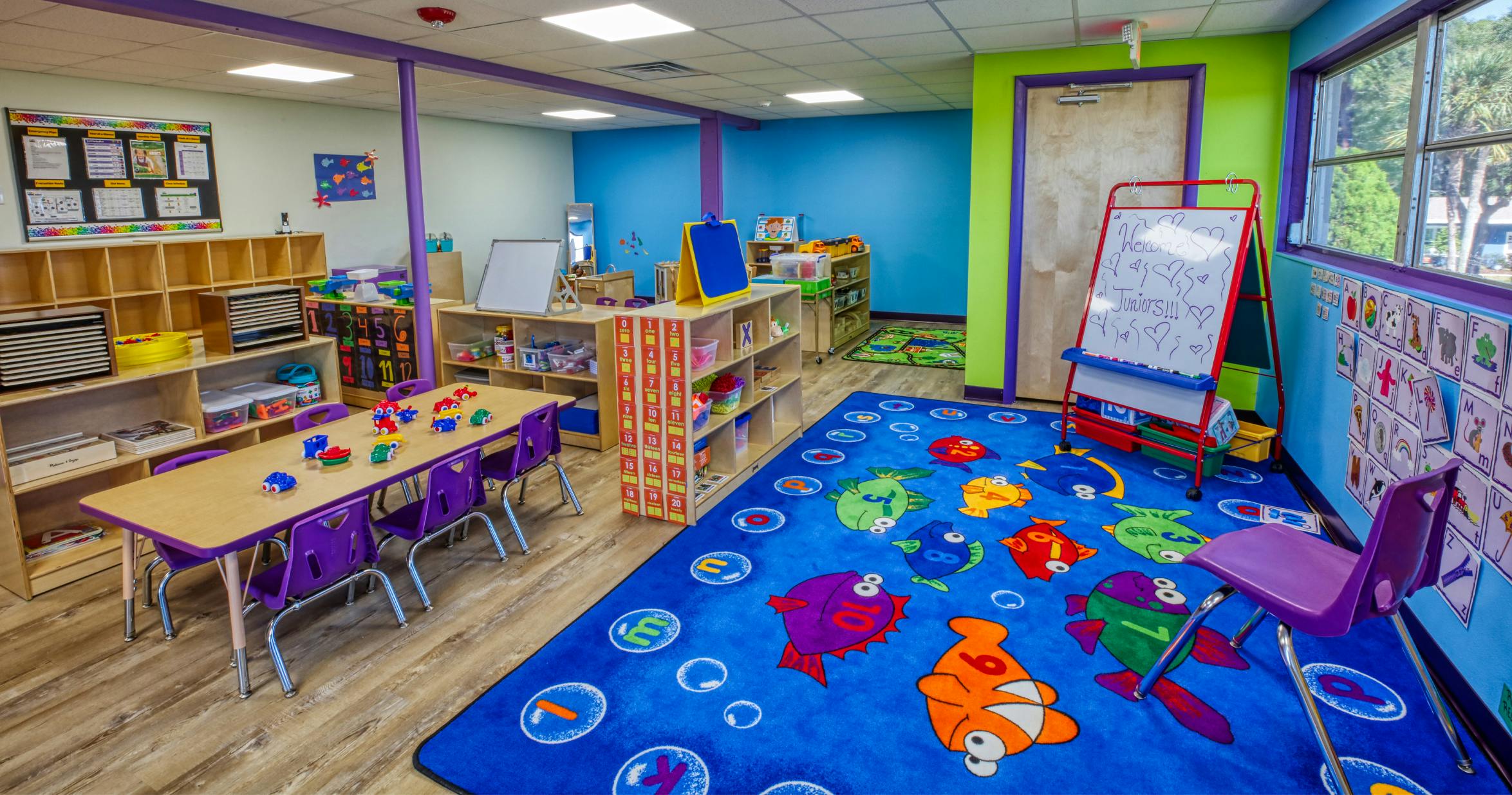 Colorful classroom with child-sized tables and chairs, shelves, and a blue rug with fish