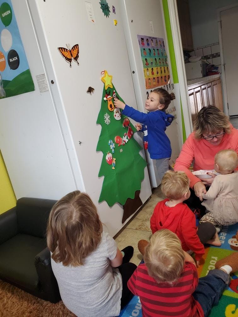 Child decorating a felt Christmas tree on a wall, with other children and an adult nearby
