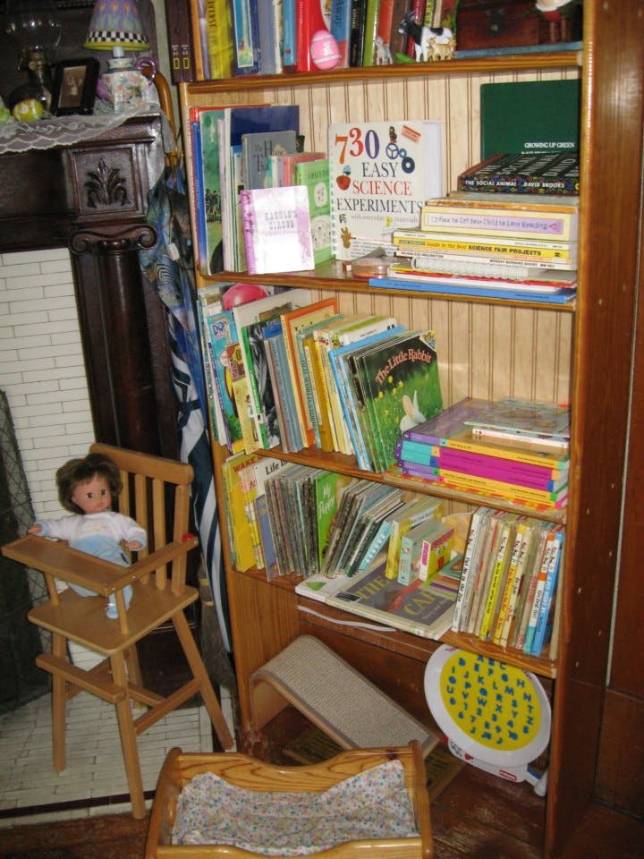 Doll in a high chair next to a wooden bookshelf filled with books