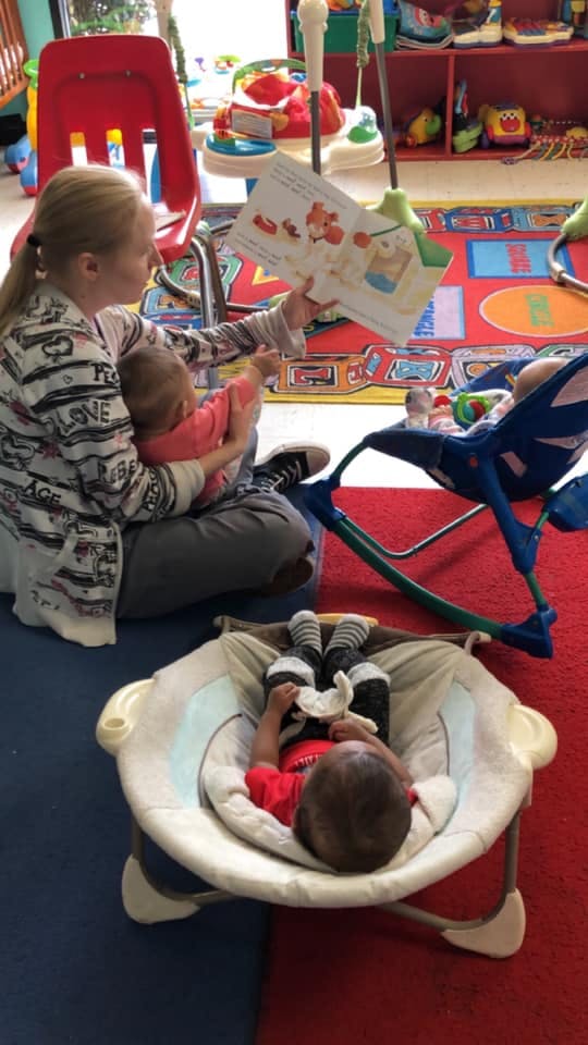 Person reading a book to a child, with another child in a bouncer