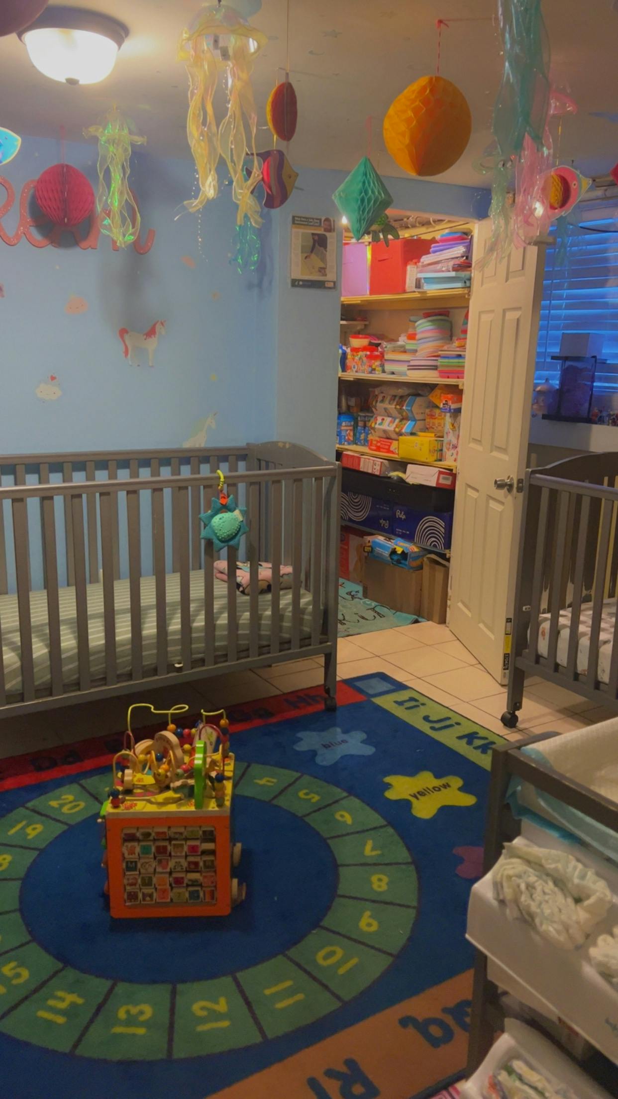 Two cribs in a room with a colorful rug and hanging decorations