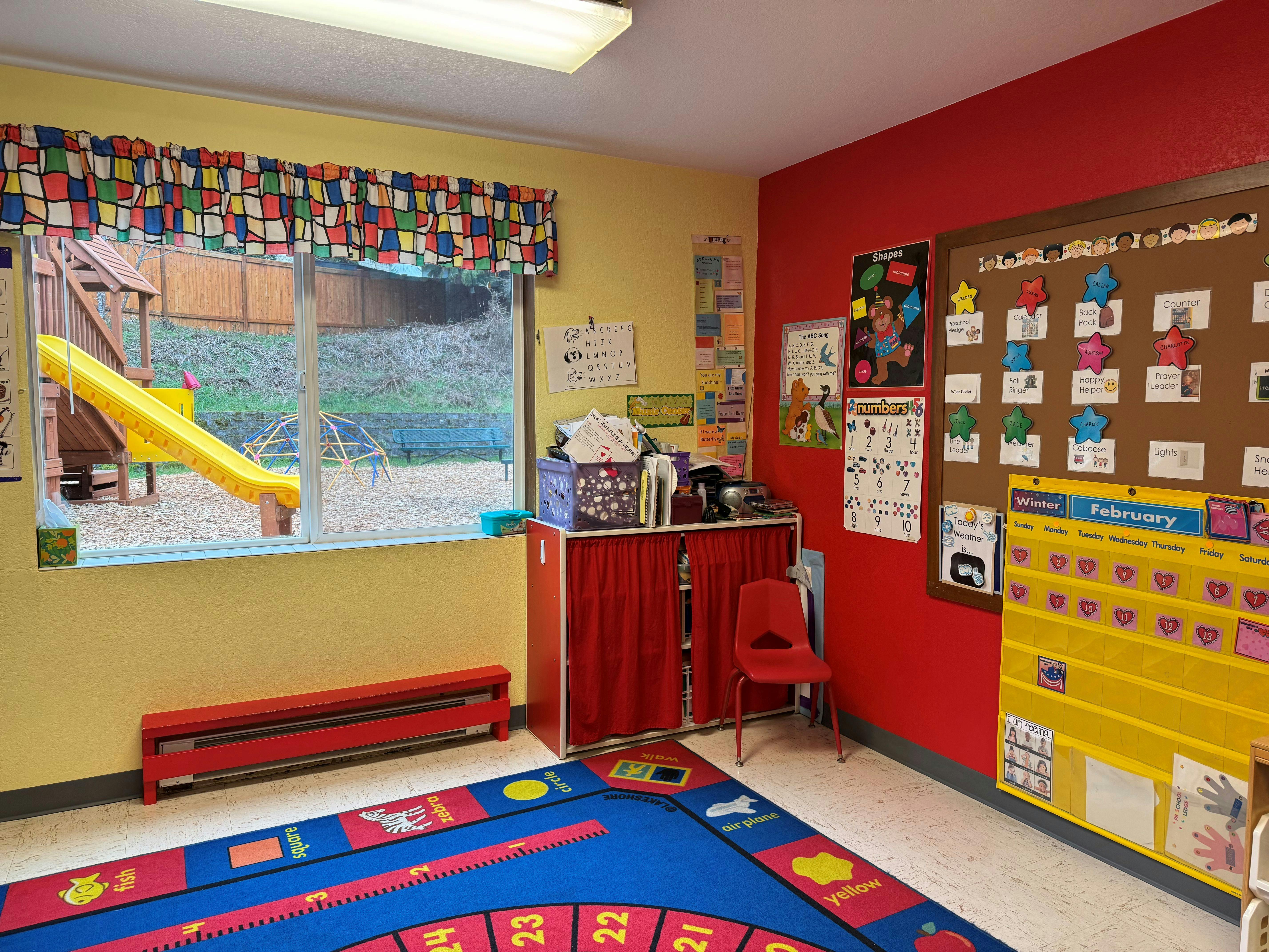 Classroom with colorful rug, calendar, bulletin boards, and view of outdoor play structure