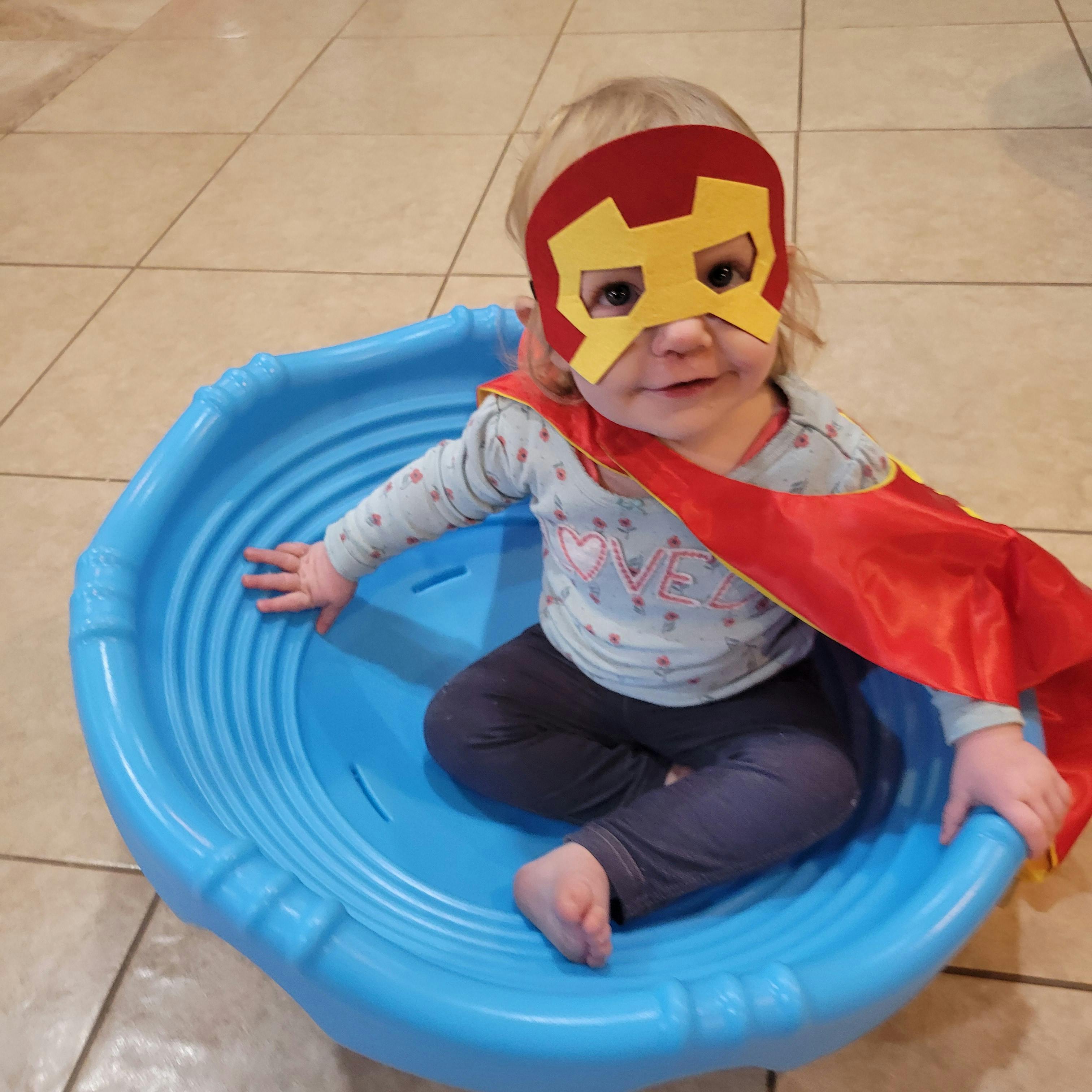 Child in a superhero mask and cape sitting in a blue plastic saucer