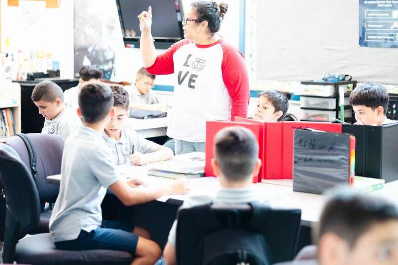 A teacher with arm raised leading a classroom of children seated at desks