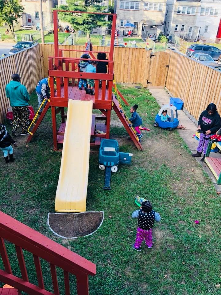 People and children playing on a red and yellow playground set in a fenced yard