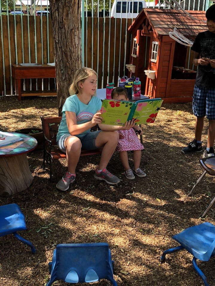 Teen and child sitting on a bench reading a book together outdoors