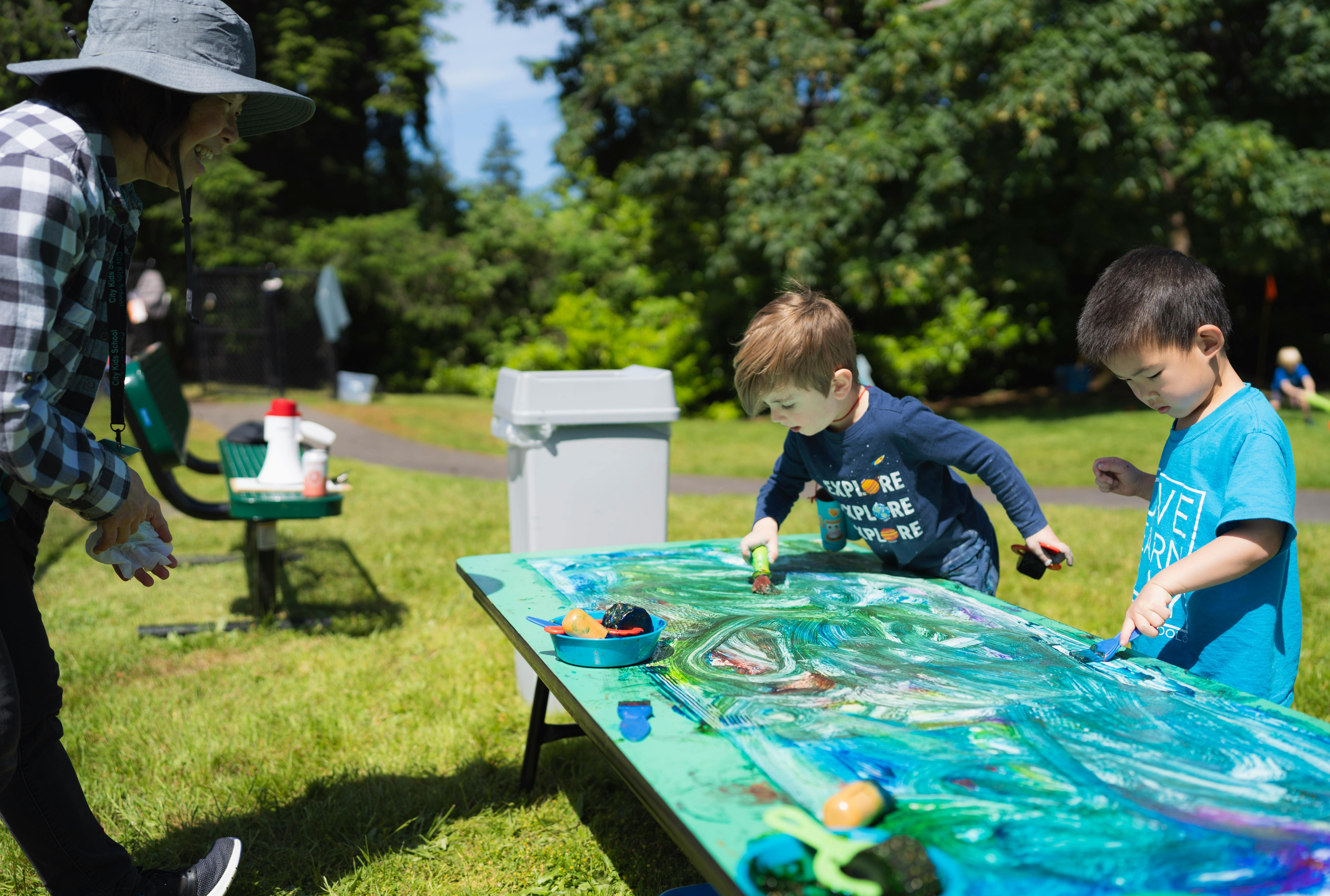 Person in hat watching two children paint on a table outdoors