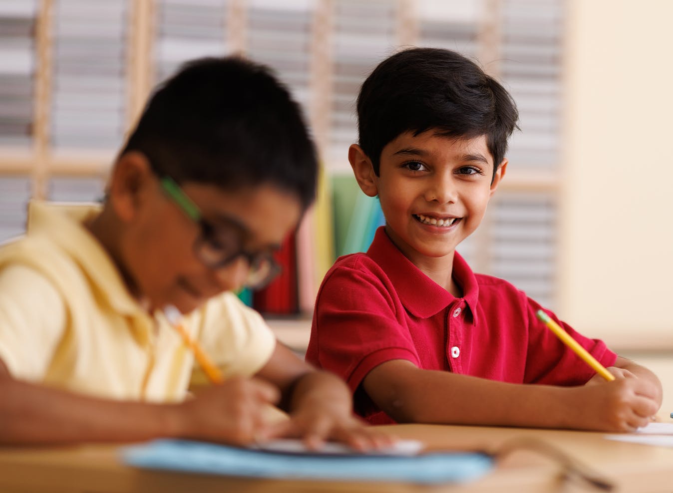 Child in red shirt smiling at camera while another child writes in background