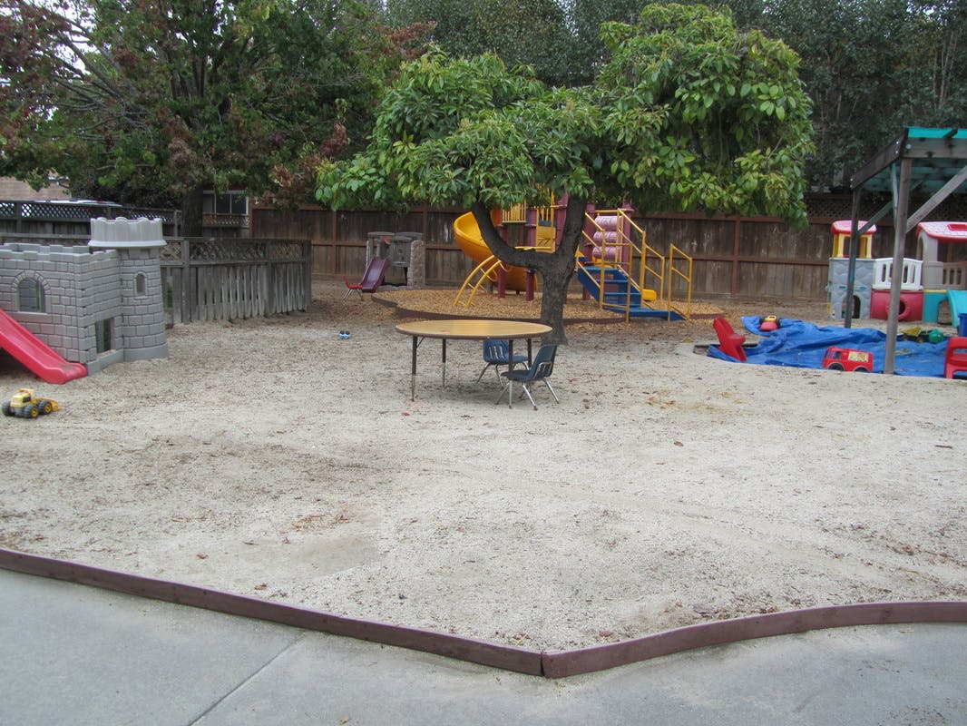 Outdoor playground with sand, play structures, and a table under a tree