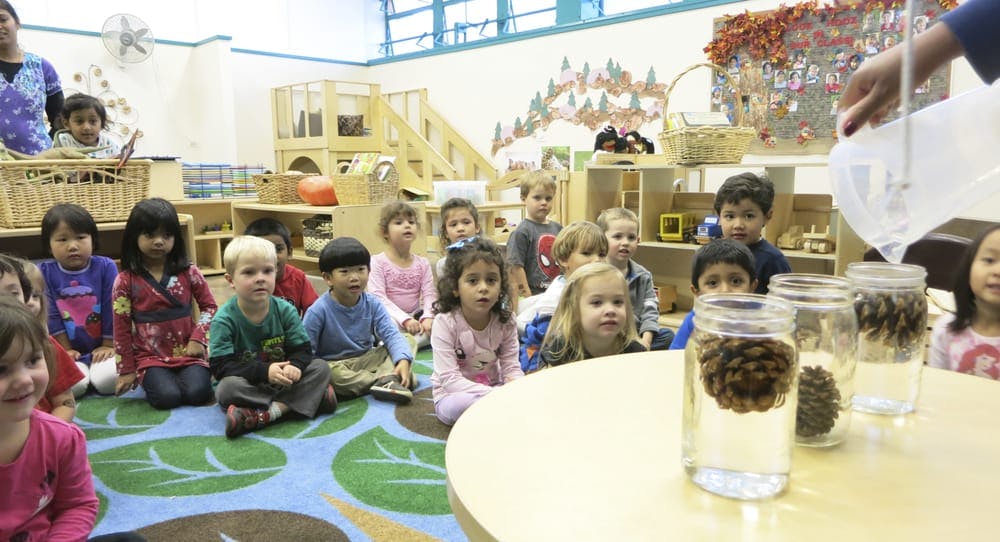 Children sitting on a rug watching a person pour water into jars