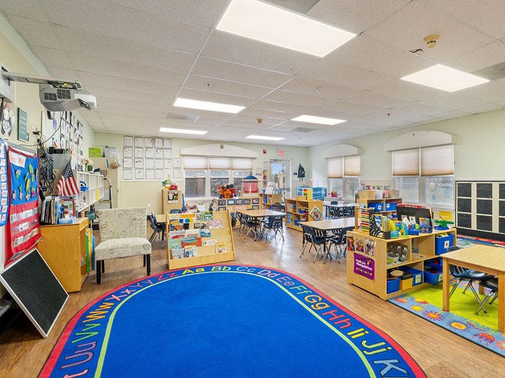 Classroom with small tables and chairs, shelves of books and toys, and an alphabet rug