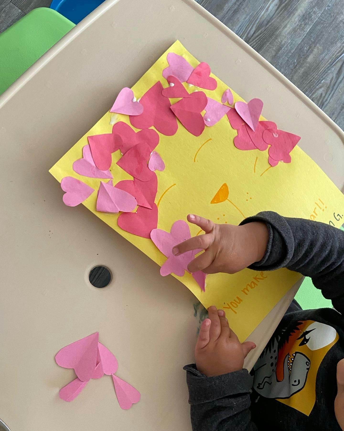 Child's hands on a yellow paper craft with pink heart cutouts