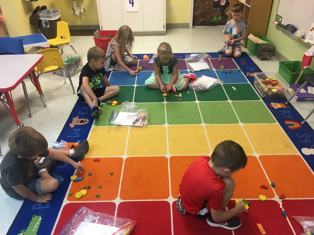 Children playing with small objects on a colorful grid rug in a classroom