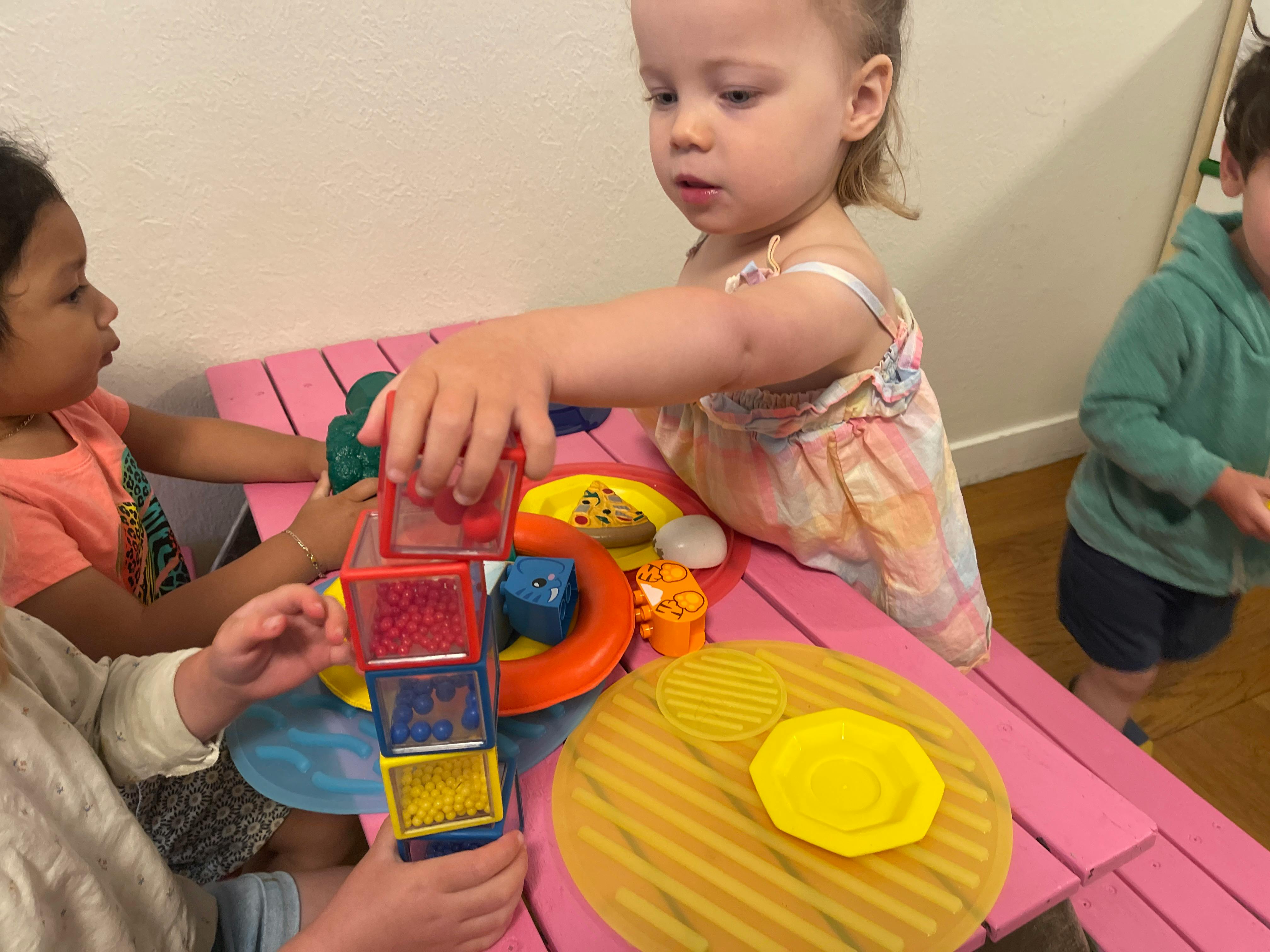 Child adding a red block to a stack of colorful sensory blocks on a pink table