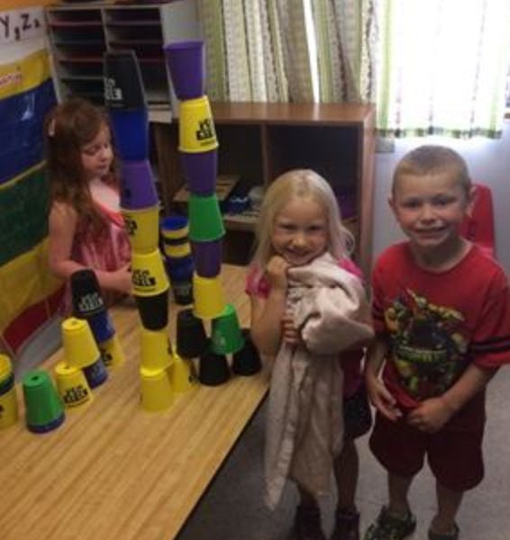 Three children standing around stacked cups in a classroom