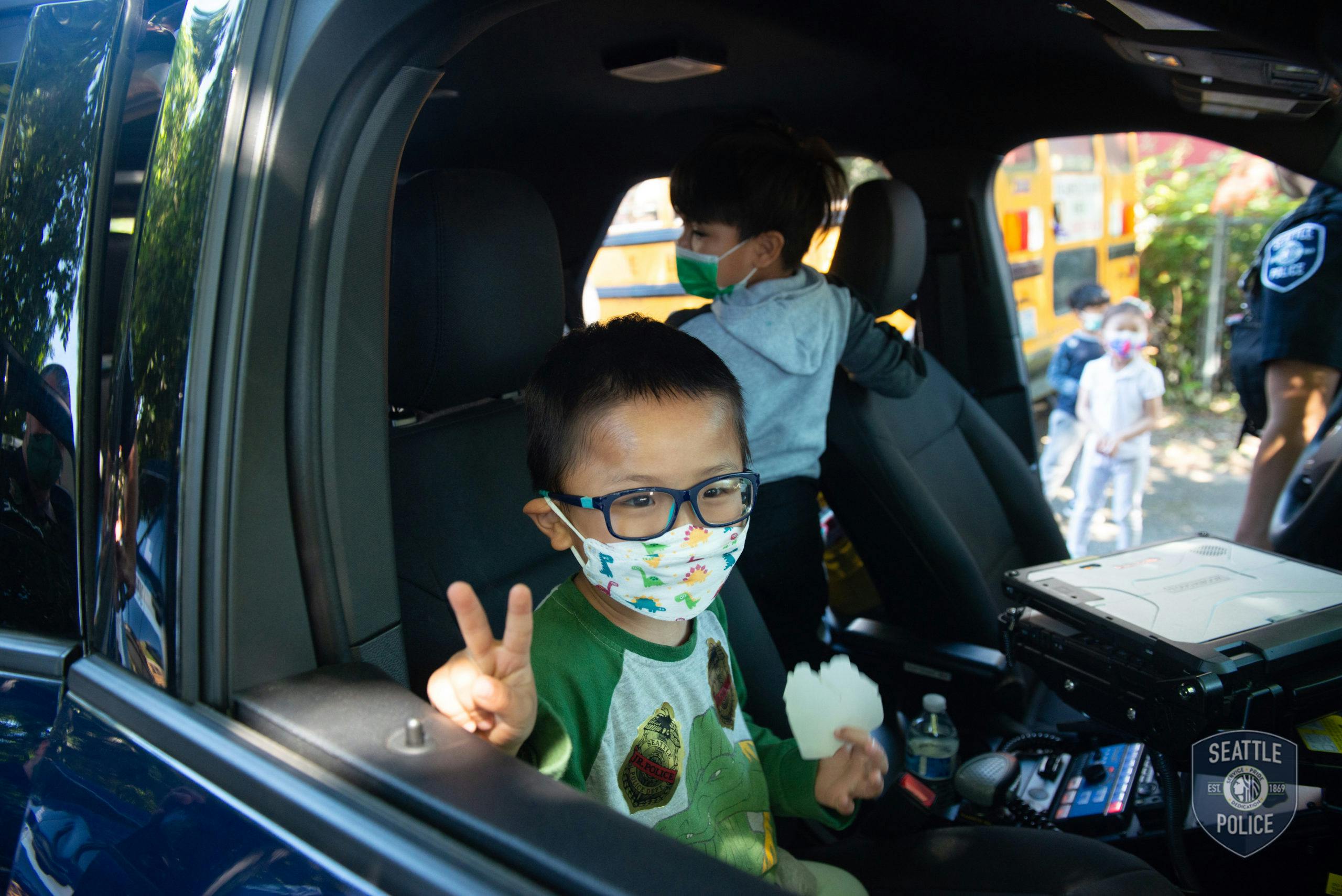 Children in masks sitting in a police car, one holding up a peace sign