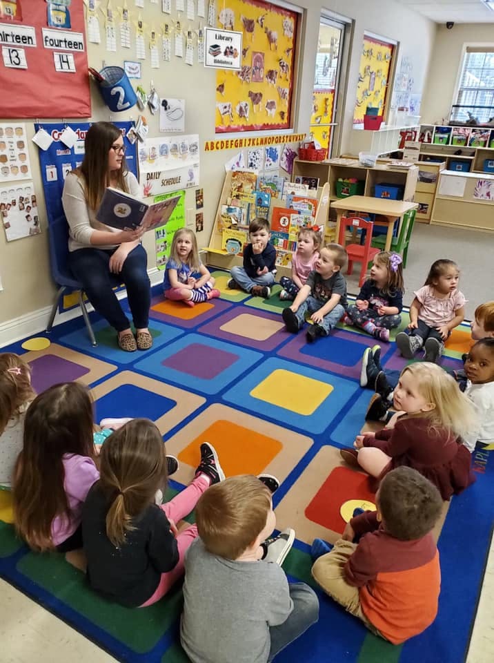 Adult reading a book to a group of children sitting on a colorful rug in a classroom