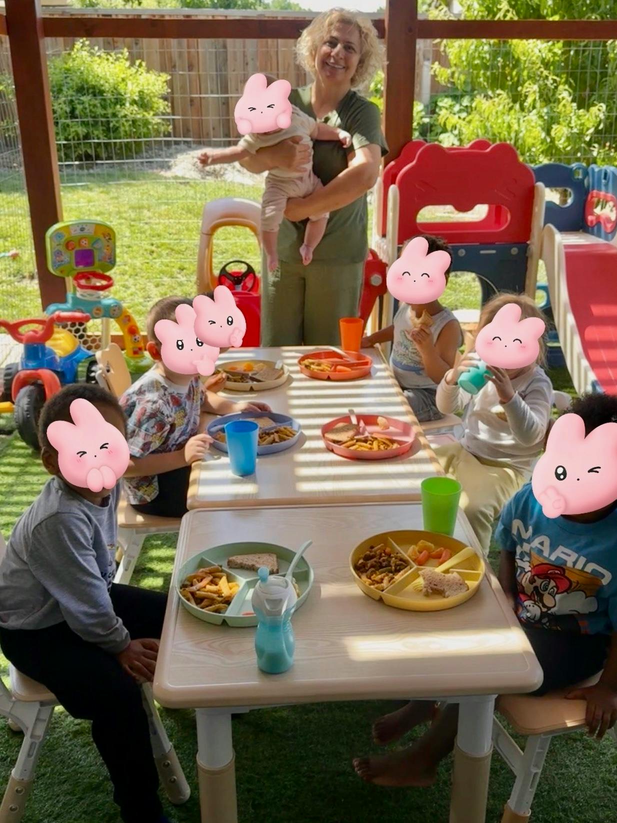 Adult holding an infant and five children sitting at tables with plates of food and drinks